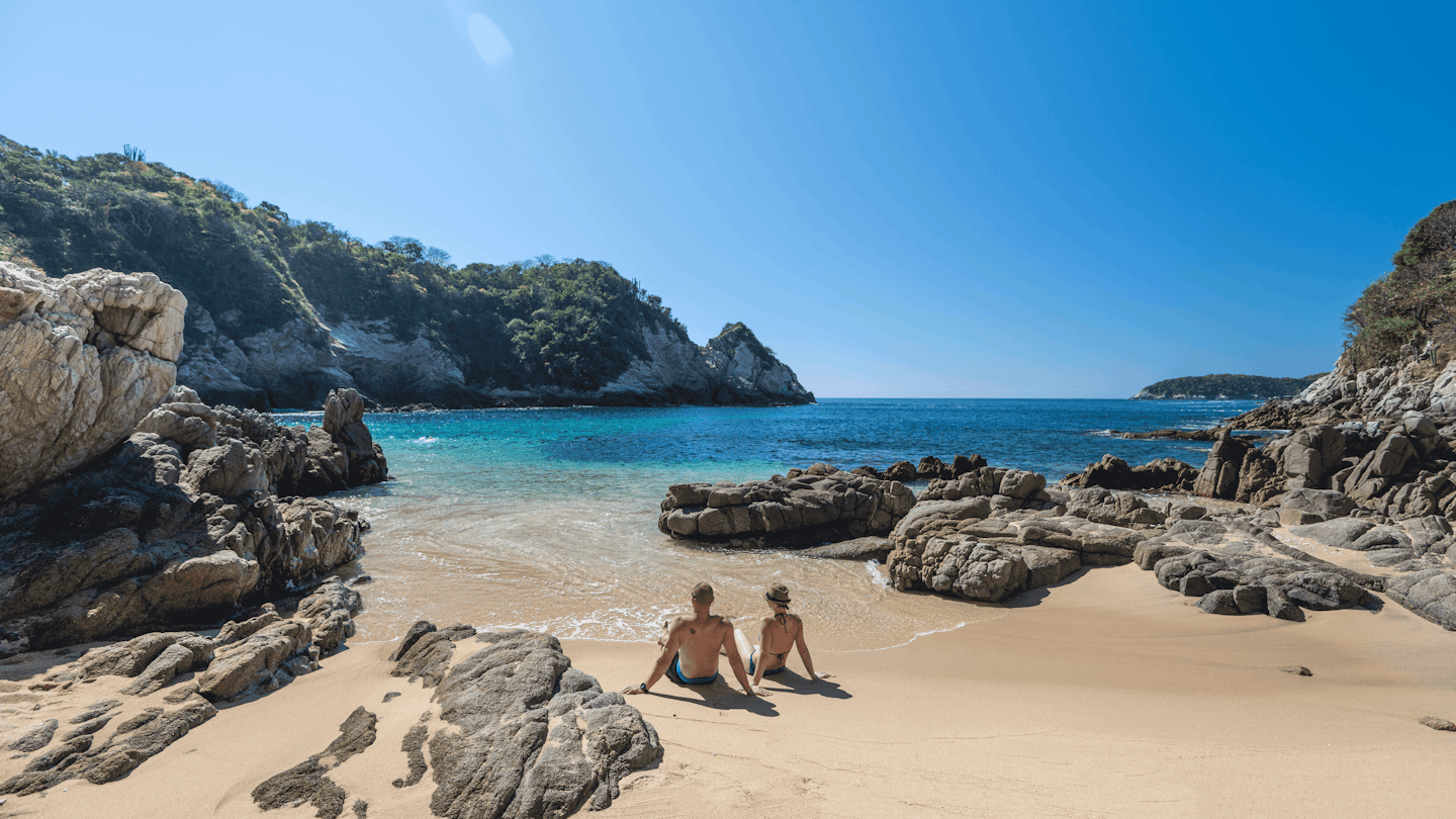A couple lounges in a sandy cove surrounded by cliffs © Ascent / Getty Images
