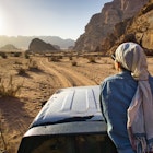 A woman tourist sitting on a car and admiring the sun light in an opening between two hills in the desert, during day time, in Wadi Rum, Jordan.