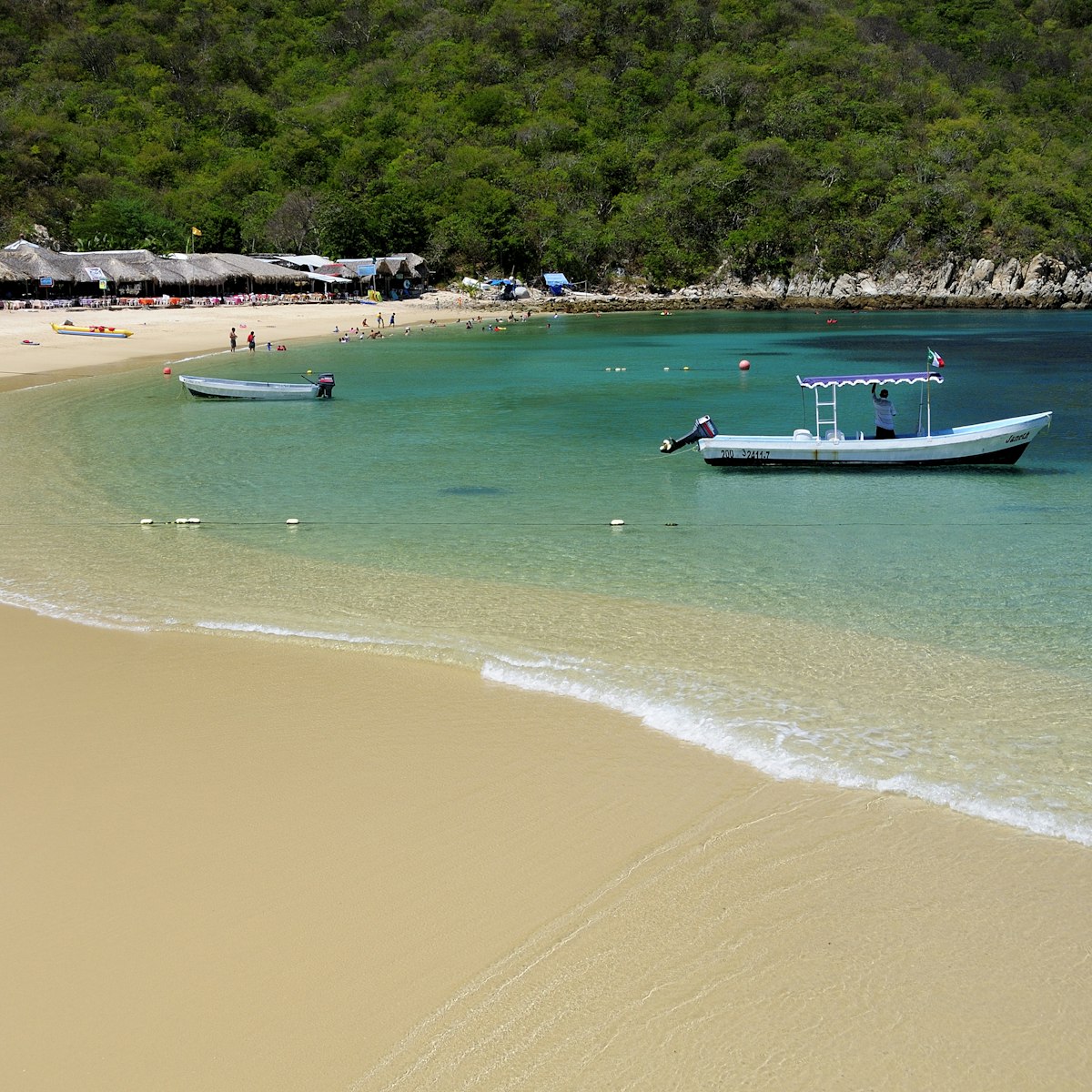Mexico, Oaxaca, Huatulco, Playa La Entrega, Stretch of sandy beach lined with thatched open fronted restaurants, with tour boat in shallow water in foreground.