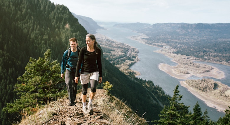 A fit older adult couple in their 50's hike up a rocky trail on a mountain ridge, the beautiful Columbia river gorge spreading out behind them. They smile as they continue up the ascent. Horizontal with copy space.
493237716
Trail, Couple - Relationship, Climbing, Outdoor Pursuit, Mature Women, Women, Men, Two People, Beauty In Nature, Columbia River, 50-59 Years, Mature Adult, Backpack, Smiling, Moving Up, Exercising, Columbia River Gorge, Healthy Lifestyle, Majestic, Discovery, Happiness, Exploration, Nature, Lifestyles, Content, Cheerful, Hiking, Gray Hair, People, Oregon, Tree, Mountain Ridge, Mountain, Attractive Person
A woman and man hiking in the hills above Columbia River Gorge