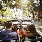 A man drives a convertible car while a woman rides in the passenger seat down a street lined with Jacaranda trees on their way out of town for a day trip from Mexico City