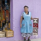 Smiling indigenous Mexican woman displaying a handwoven Zapotec rug outside a shop in Santa Ana del Valle.