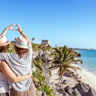 two women with their arms around each other create a heart with their hands while looking at a Mayan ruin on the beach