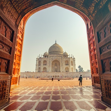 The Taj Mahal as viewed through the doorway of the main gate, which frames the building neatly. Crowds walk towards the white marble structure, which has a domed roof and four pillars surrounding it.