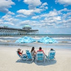 Family relaxing on the beautiful beach, People enjoying summer vacation by the ocean. Family sitting under beach umbrella. Cloudy sky and pier in the background. Folly Beach, South Carolina USA. ; Shutterstock ID 1428607988; your: Sloane Tucker; gl: 65050; netsuite: Online Editorial; full: Free Things South Carolina Article